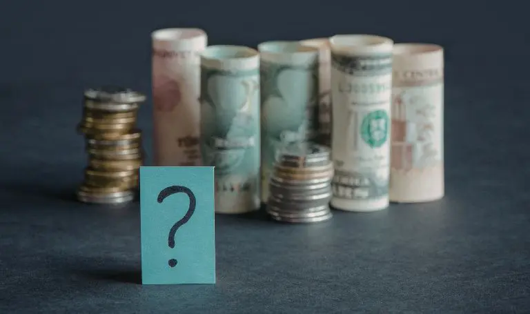 Rolls of paper currency and stacks of coins behind a teal card with a question mark, symbolizing uncertainty about business cash