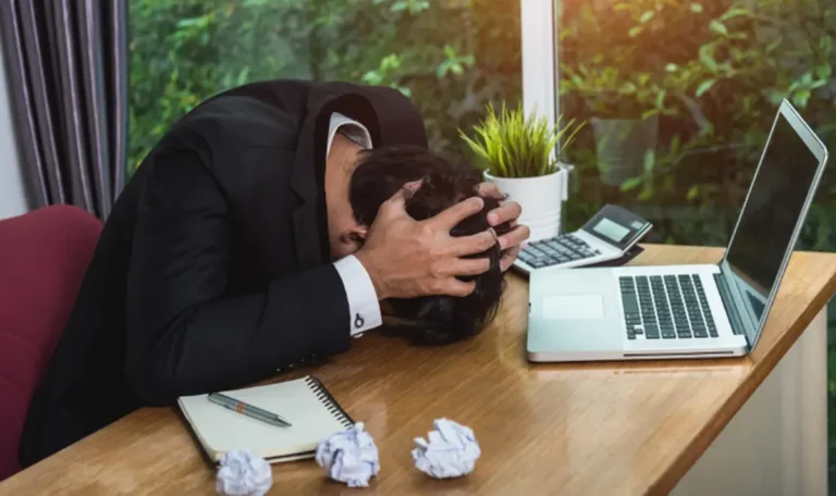 Stressed business owner in a suit with head in hands at a wooden desk, surrounded by crumpled paper, a laptop, and a calculator